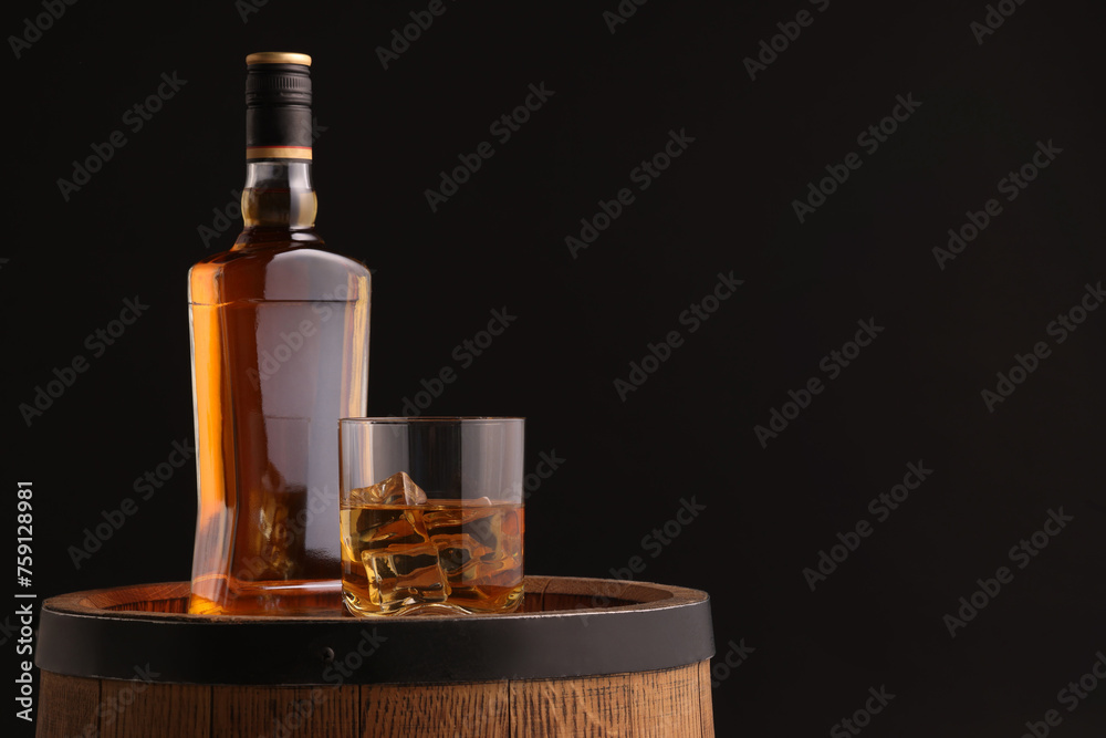 Whiskey with ice cubes in glass and bottle on wooden barrel against black background, space for text Whiskey with ice cubes in glass and bottle on wooden barrel against black background, space for text