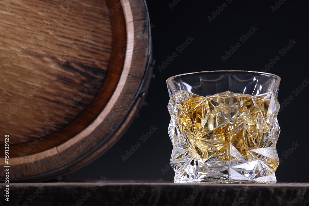 Whiskey with ice cubes in glass and barrel on wooden table against black background, closeup. Space for text