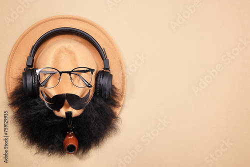 Man's face made of artificial mustache, beard, glasses and hat on beige background, top view. Space for text