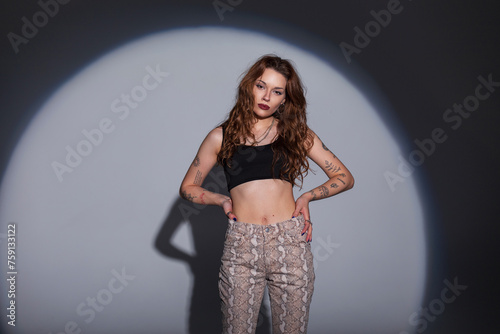 Cool beautiful hipster woman with tattoos on her arms in a fashion black top with jeans with a snake print stands on a dark background in the studio