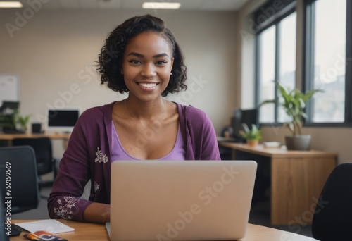 A woman in a purple cardigan smiles at her laptop in a bright office. She appears content and focused. photo