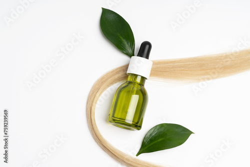 A strand of soft blonde hair encircles a green glass dropper bottle, paired with lush green leaves, highlighting organic haircare oils photo