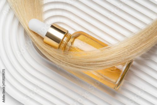A luxurious golden hair oil in a clear dropper bottle rests on blonde hair against a wavy white background, symbolizing upscale hair care