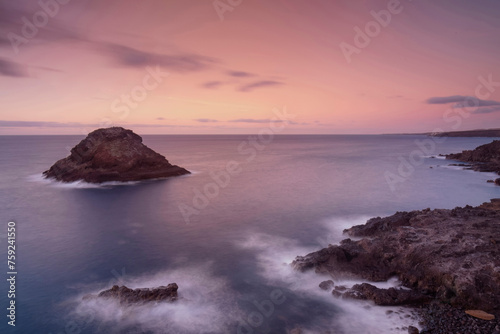A pastel sunset sky softens the seascape around a rocky islet off the rugged coastline, creating a dreamlike vista photo