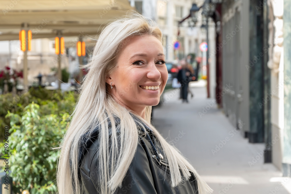 A blonde 35-40 year old woman stands in a blurred background of a European street and smiles.