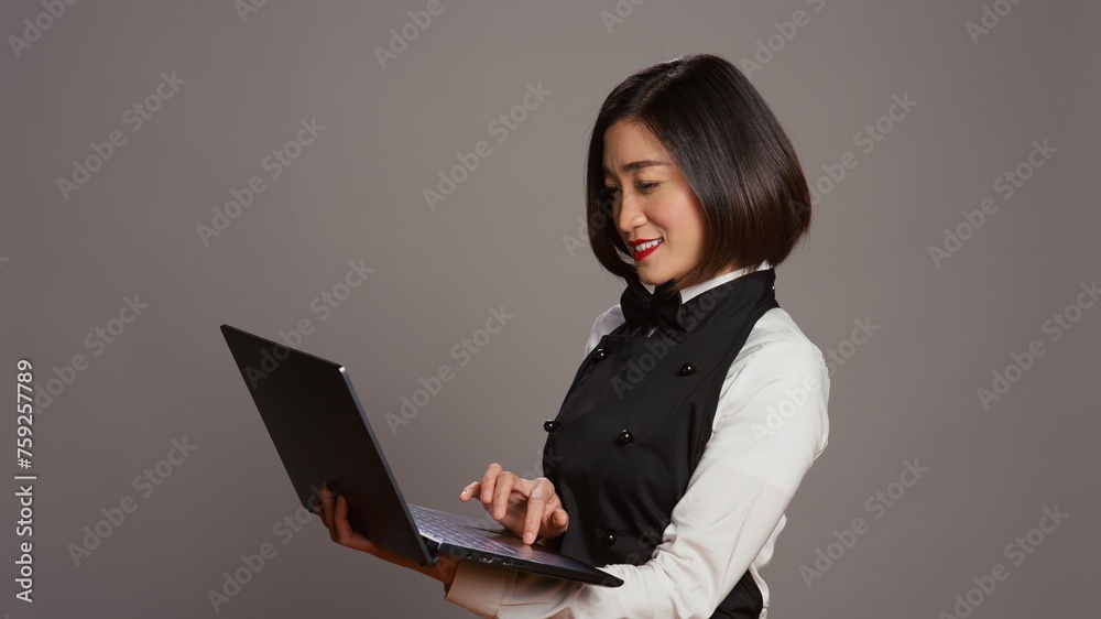 Asian waitress checking all table reservations online on laptop, creating a list of customers and menus at restaurant. Catering employee with apron using pc to offer great service. Camera B.