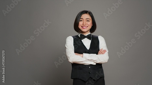 Asian woman hotel concierge wearing elegant uniform with bow, working in hospitality industry as a receptionist. Reception desk administrator greeting clients, grey background. Camera A.