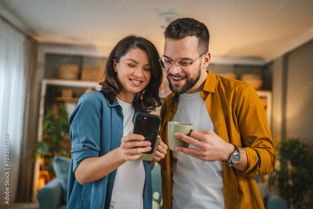 © Miljan Živković - Adult couple man and woman husband wife have a cup of coffee use mobile phone search internet social network happy smile