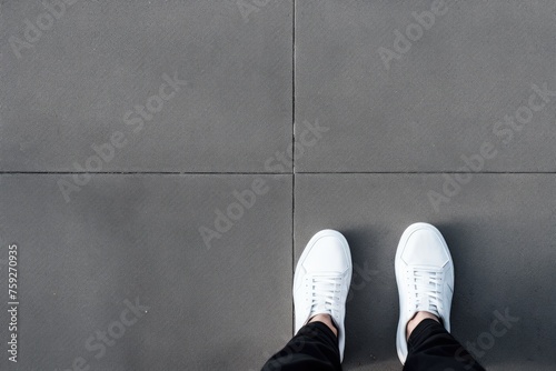 A person's feet clad in clean white sneakers, positioned on a textured grey pavement. White Sneakers Standing on Grey Pavement © Anatolii