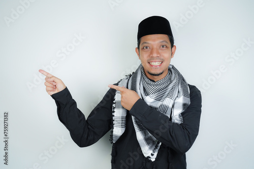 Cheerful handsome Asian Muslim man wearing Arab turban sorban pointing hand finger at empty space for advertising on isolated background photo