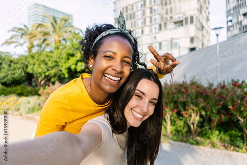 Two young beautiful women taking selfie portrait together in summer at city. Diverse girls enjoying free time outdoors. Female friendship concept.