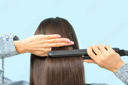 Beautiful young woman straightening hair on blue background, back view