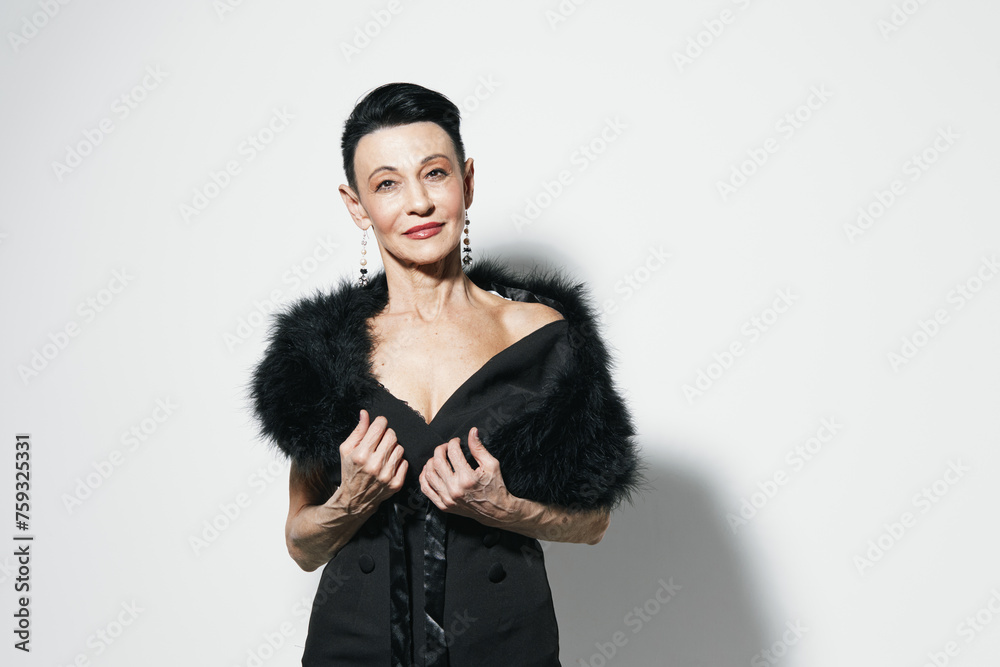 Fashionable woman in elegant black dress and fur stole posing confidently against white wall with hands on hips Fashionable woman in elegant black dress and fur stole posing confidently against white wall with hands on hips