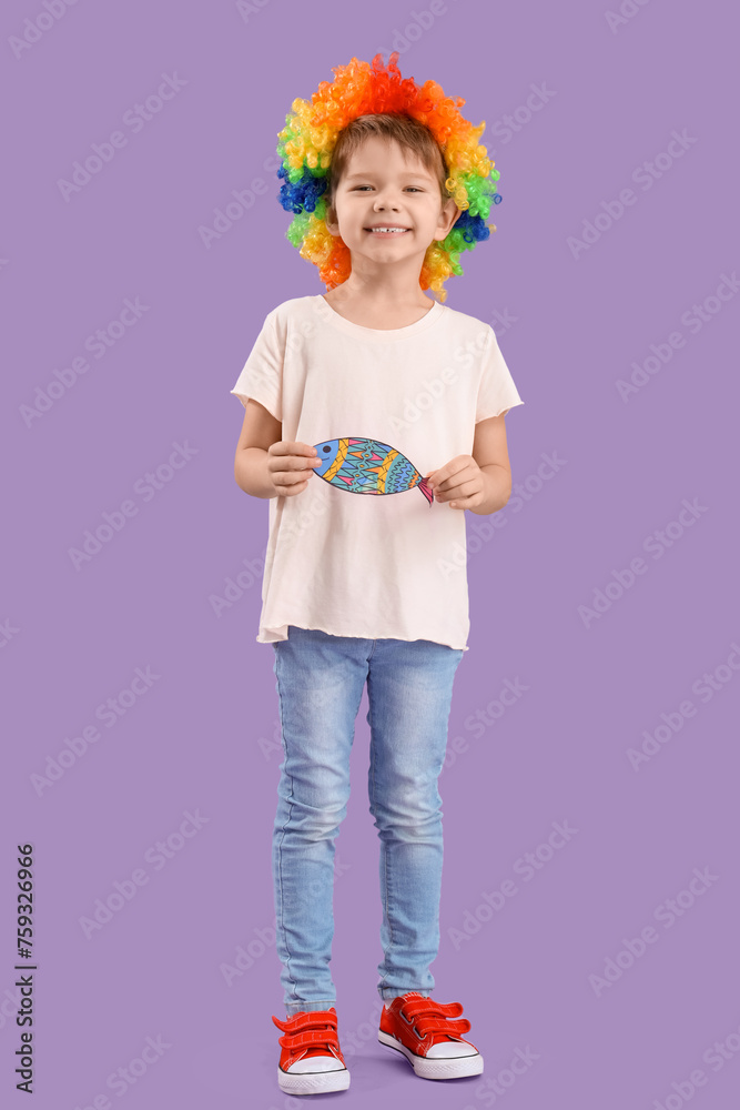 Cute little boy with colorful clown wig and paper fish on lilac background. April Fool's Day celebration