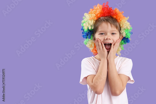 Happy little boy with colorful clown wig on lilac background. April Fool's Day celebration