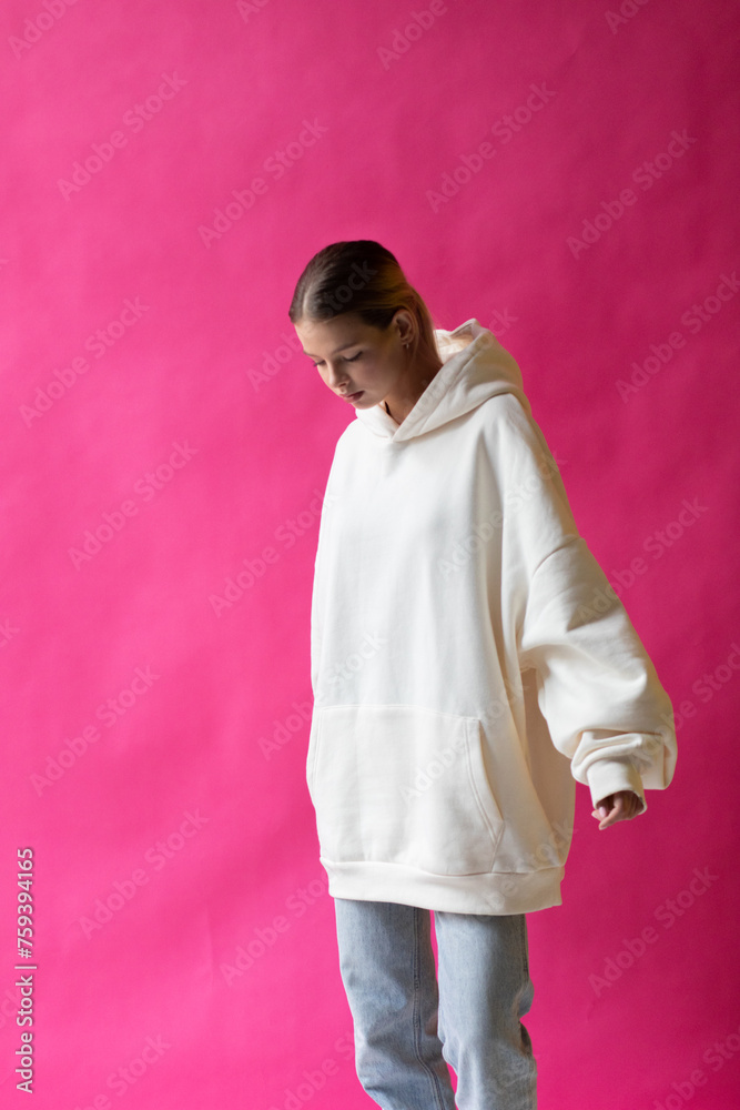 Fototapeta premium Beautiful blonde woman in a white hoodie and blue jeans posing on a pink background