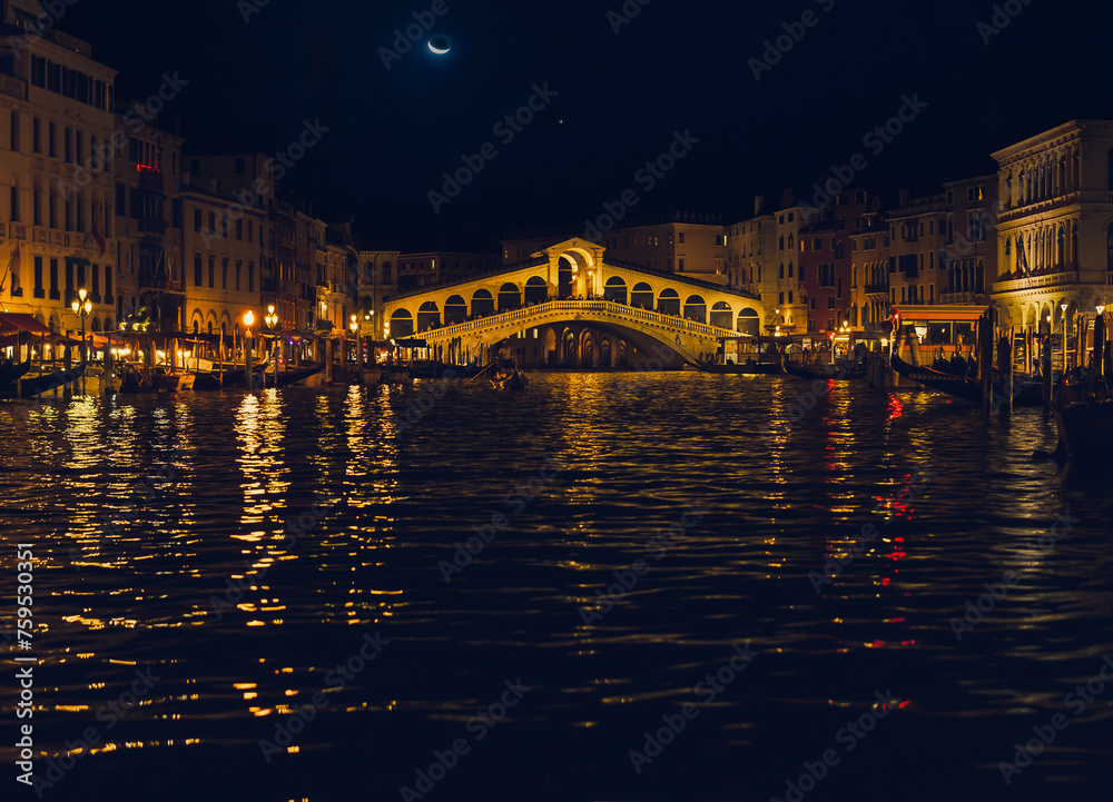 View of Rialto bridge in Venice, Italy, with crescent moon late in evening; city lights reflect in dark water