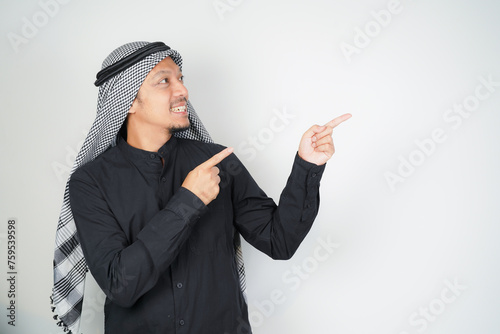 Cheerful Asian Muslim man wearing Arab turban sorban pointing hand finger at empty space on isolated photo