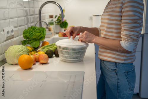 Slim vegan woman preparing useful dinner of fresh fruit and vegetables. Concentrated female drying greenery in plastic hand food centrifuge. Tasty ripe oranges, apples. cabbage, avocado lying on table