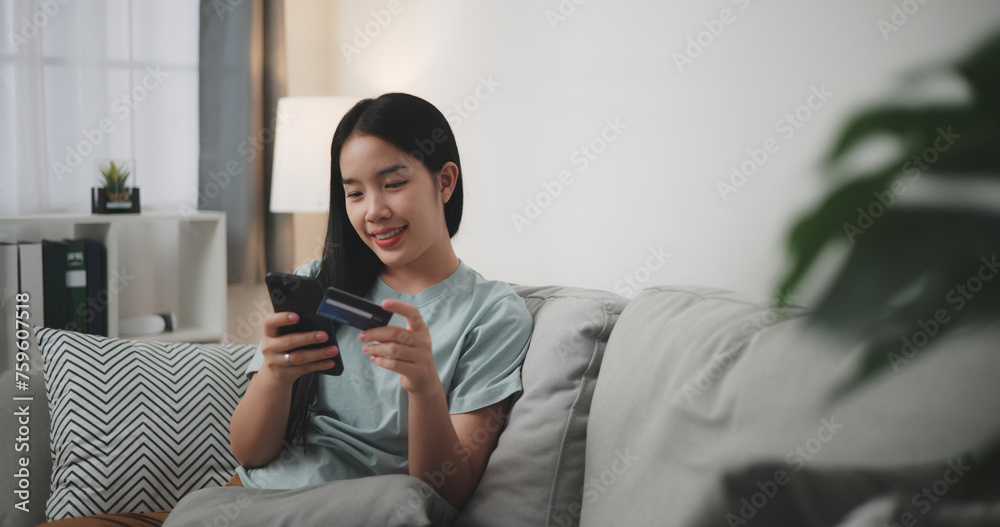 Selective focus, Young asian woman sitting on sofa holding credit card making online payment for purchase in web store using smartphone. Selective focus, Young asian woman sitting on sofa holding credit card making online payment for purchase in web store using smartphone.