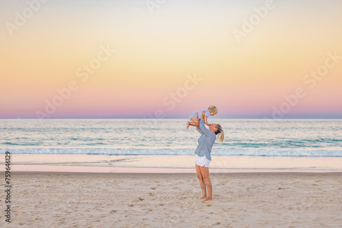 Mother lifting toddler son in the air on the beach with pink sunset sky photo