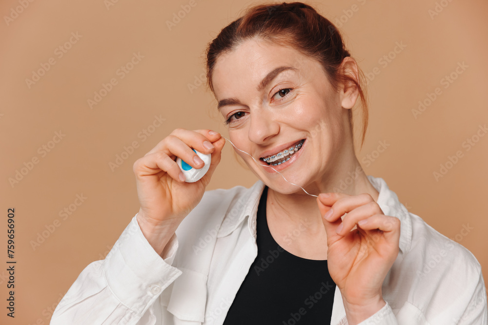 Portrait of mature woman in braces holding dental floss near her teeth on beige background Portrait of mature woman in braces holding dental floss near her teeth on beige background