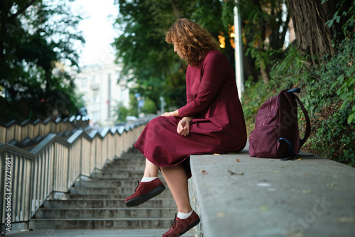 Cheerful young caucasian woman smiles with her teeth looking to side, sitting in European city on pavement. Brunette wears dress. City life concept. High quality photo