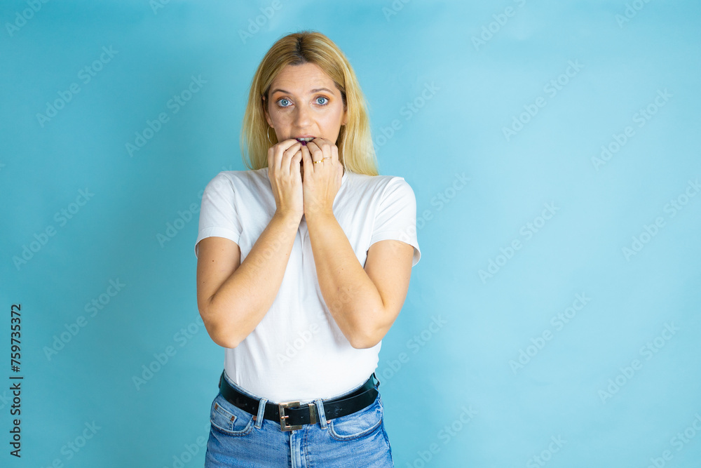 Young beautiful woman wearing casual t-shirt over isolated blue background looking stressed and nervous with hands on mouth biting nails. anxiety problem. Young beautiful woman wearing casual t-shirt over isolated blue background looking stressed and nervous with hands on mouth biting nails. anxiety problem.