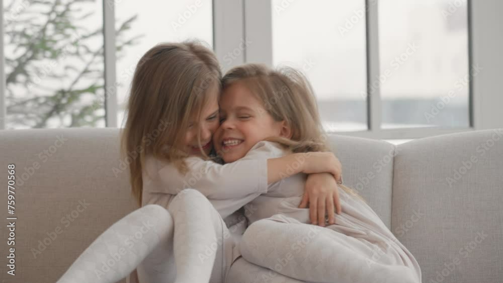 Two children twins playing at home. Happy smiling mischievous little sisters twins sitting on sofa, hugging and laugh, playing in living room at home. Family having fun, sisters' friendship concept.