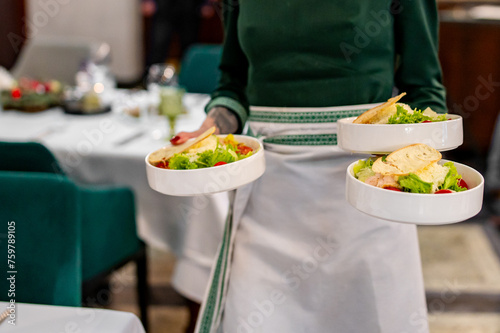 A waiter serves fresh salads in an elegant dining setting. The scene features white bowls filled with green lettuce, vegetables, and garnishes. Soft lighting creates a warm ambiance