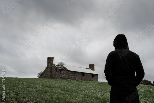 A mysterious hooded figure looking at an abandoned ruined farm house on a hill. On a grey stormy day in the countryside.