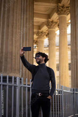 Handsome young man in sunglasses taking selfie with smartphone. Male hipster making video chat via mobile phone outside. Vertical view.