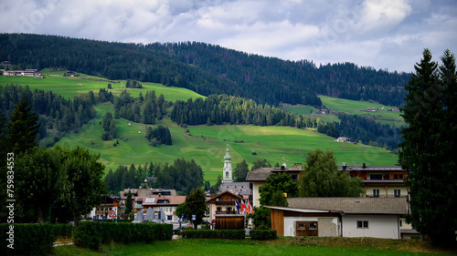 Ein Dorf mit einer Kirche im Zentrum vor einem Berghang der mit Wiesen und Wald   berzogen ist