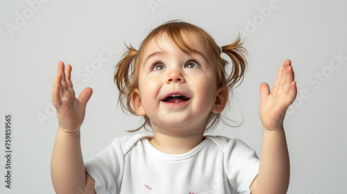 joyful toddler with pigtails is raising her hands in excitement and looking up, set against a plain light background