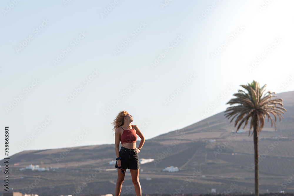 A woman stands on a hill overlooking a palm tree. She is enjoying her vacations on lanzarote. The sky is clear and the sun is shining brightly. Copy space.
