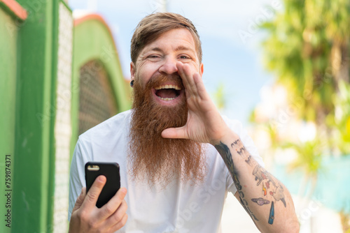 Redhead man with beard using mobile phone at outdoors shouting with mouth wide open