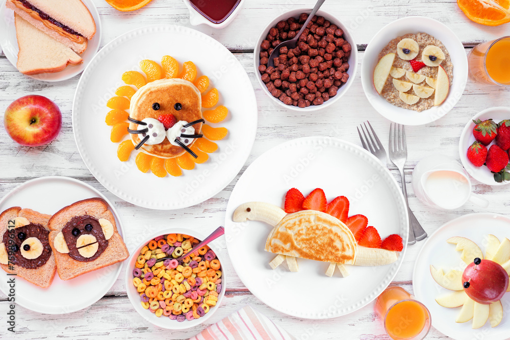 Fun child theme breakfast table scene with various animal themed foods. Top view on a white wood background. Pancakes, oatmeal, toast, fruit and cereal.