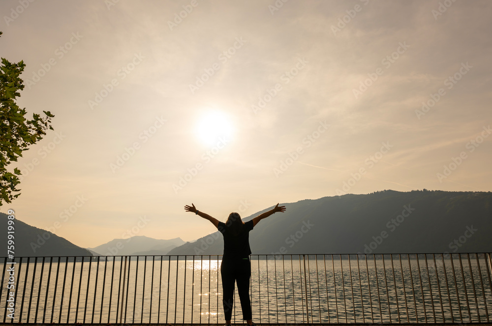 Fototapeta premium Woman Standing with Arms Outstretched on the Waterfront with Railing to Lake Lugano with Mountain in a Sunny Summer Day in Lugano, Ticino, Switzerland.