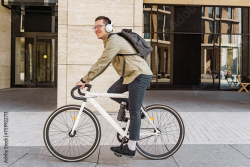 Smiling man riding a stylish white bicycle in front of a modern building, wearing headphones and a backpack.