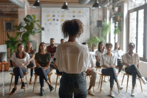 Confident female business coach teaches small group of people new business knowledge. People are sitting on chairs in office of business center listening to woman standing with her back to camera photo