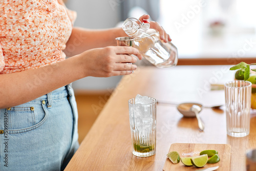 drinks and people concept - close up of woman pouring water from glass bottle to jigger and making lime mojito cocktail at home kitchen photo