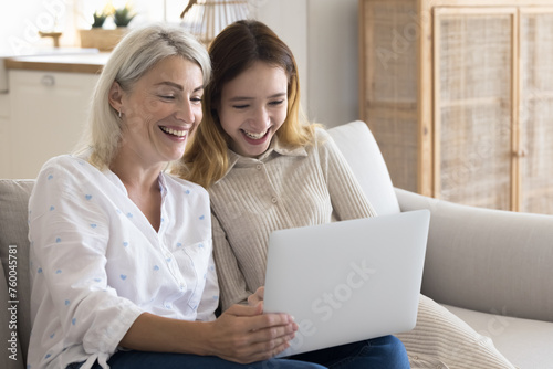 Cheerful mature mother spend time with teen daughter, sit on couch with laptop, watch funny video or movie, make purchase via web-store, learn new program, browse internet. Family leisure, technology