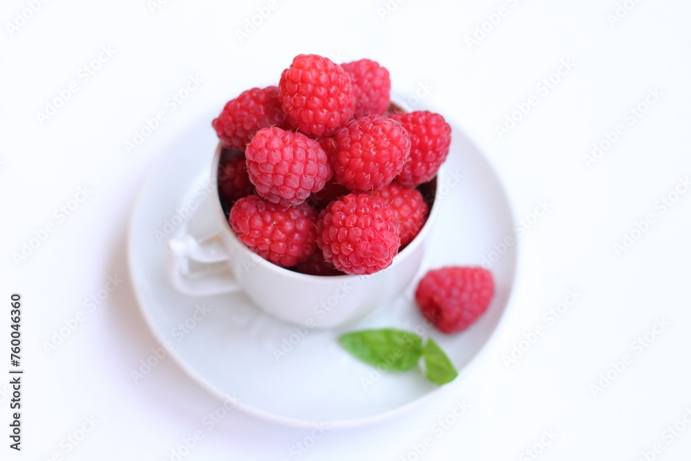 Lots of raspberries in a white rustic cup, green basil leaf on a white background isolated. Fresh ripe summer berries on a plate. Close. up Lots of raspberries in a white rustic cup, green basil leaf on a white background isolated. Fresh ripe summer berries on a plate. Close. up