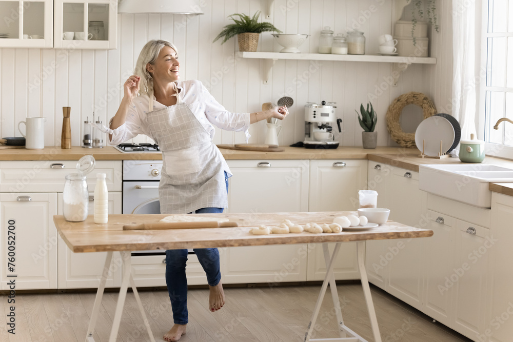 Cheerful mature woman in apron dancing in the kitchen, listen favorite music, moving, feel carefree, prepare holiday cake for family, enjoy culinary hobby alone at home. Domestic activity, cooking Cheerful mature woman in apron dancing in the kitchen, listen favorite music, moving, feel carefree, prepare holiday cake for family, enjoy culinary hobby alone at home. Domestic activity, cooking