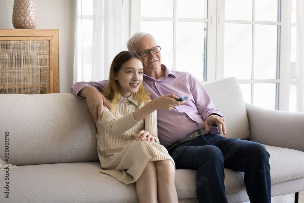 Pre-teen granddaughter holds remote control, switch channels spend leisure with older grandfather, rest, sit together on sofa. Family pastime at home, online digital streaming TV services users, tech