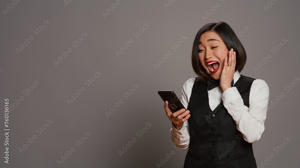 Front desk staff in uniform looks at online information on mobile phone, checking all website reservations over grey background. Asian receptionist using smartphone for bookings. Camera B.
