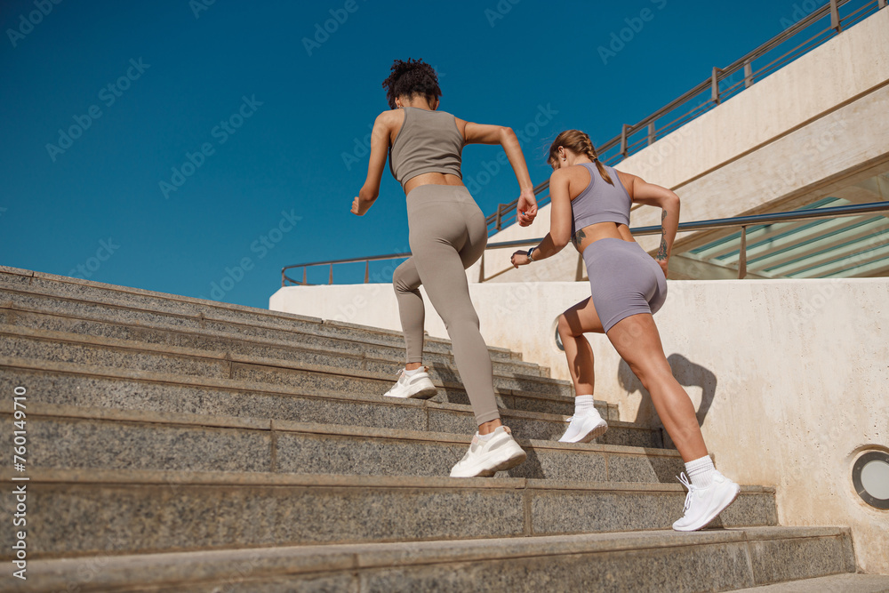 Two active female athlete friends in sportswear running on steps outdoors on a sunny day