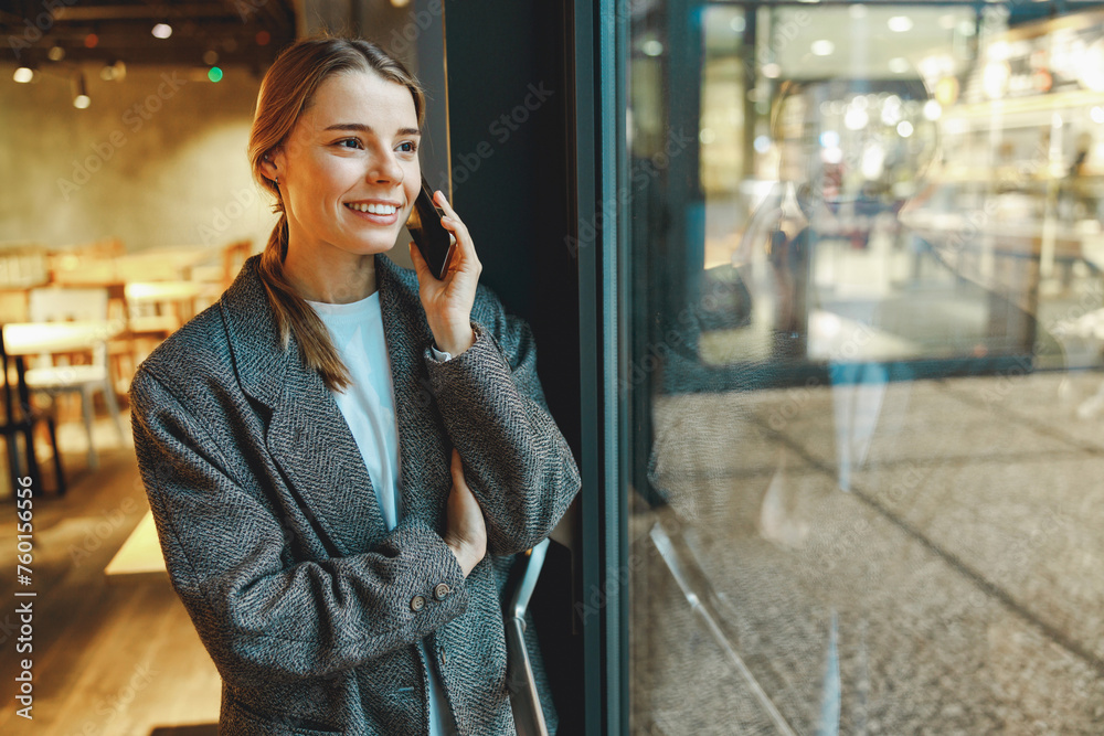 Fototapeta premium Stylish young female freelancer is talking phone with client while standing in coworking near window