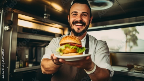 Cheerful chef in white uniform holds out a burger towards the camera in a bright kitchen