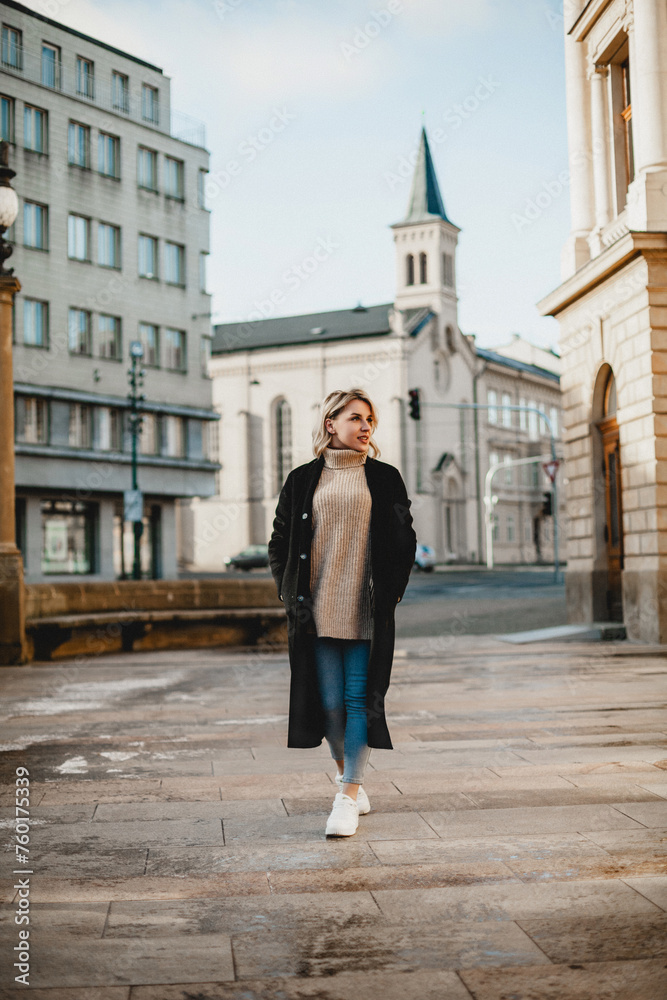 Stylish Woman in Black Coat and Blue Jeans Strolling Through Urban Landscape. Moody Cityscape with Fashionable Female Walking Along City Street. Urban Chic Concept with Woman Amidst City Buildings.